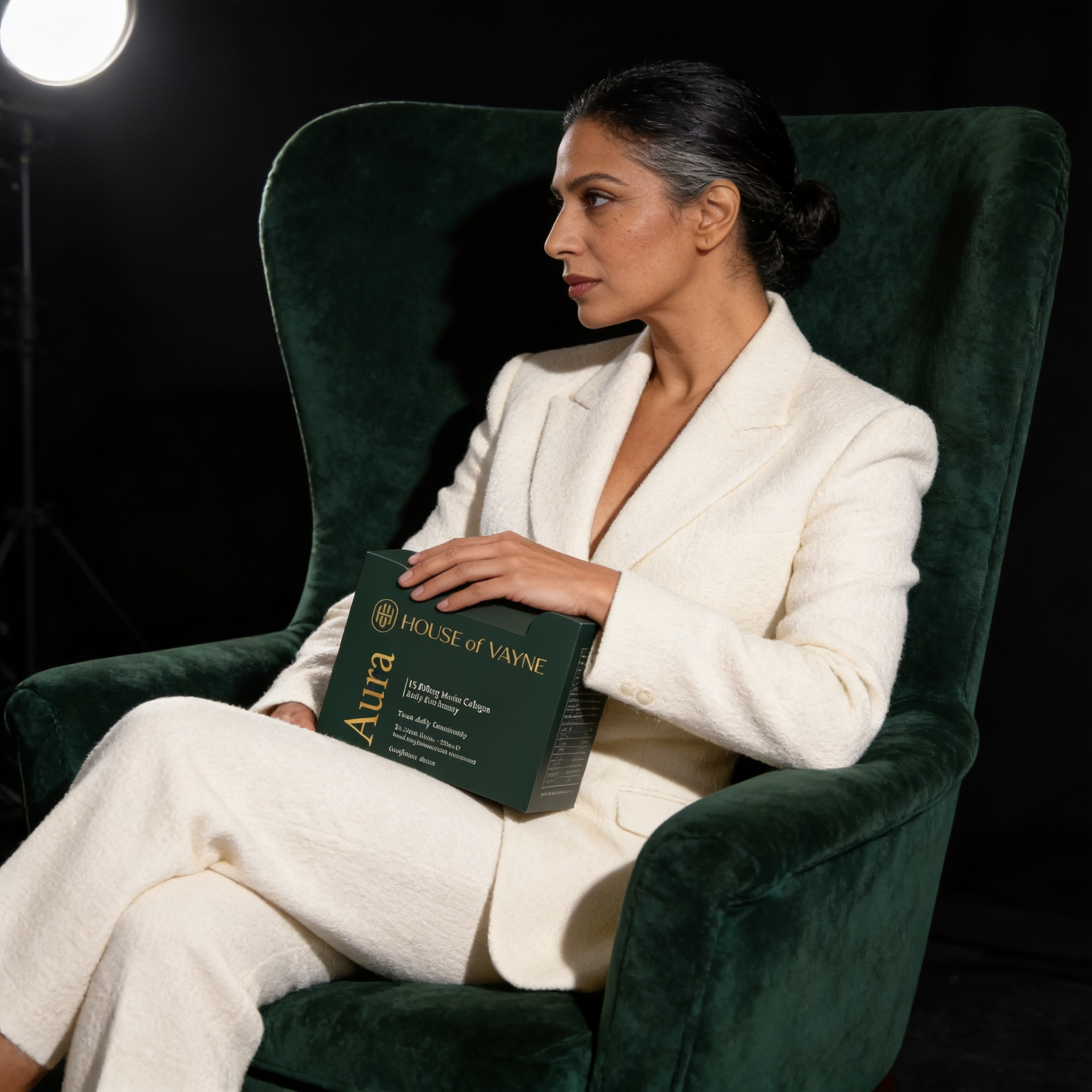 Mature woman in cream tailored suit seated on dark green velvet chair holding Aura box